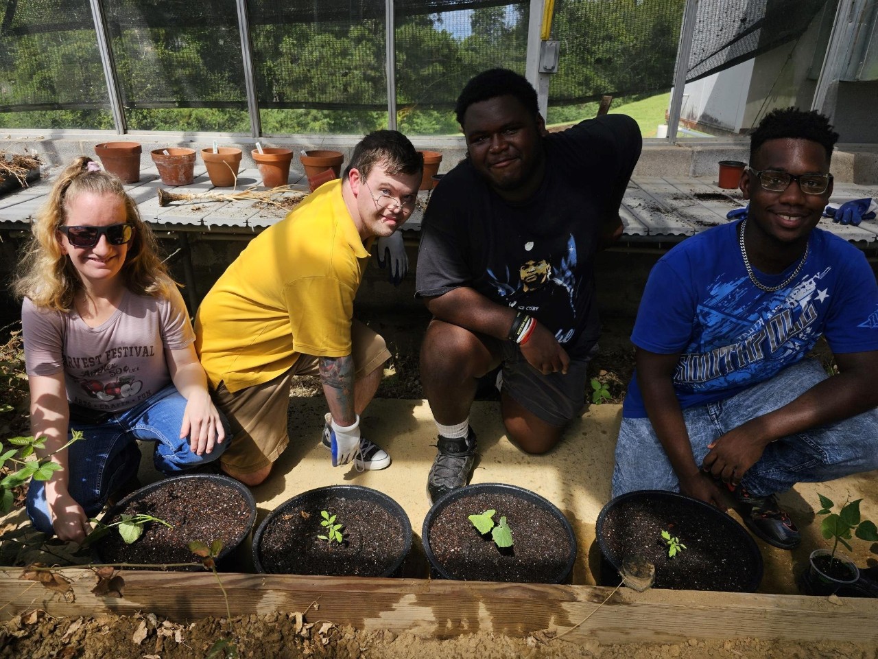 Students in greenhouse Image Description: Four people in a greenhouse setting, all smiling and engaged in plant care activities. From left to right: 1. A light-skinned woman with blonde hair wearing sunglasses, a light-colored t-shirt with a graphic print, and blue jeans. 2. A light-skinned man with short brown hair, glasses, and a yellow t-shirt, crouched down and smiling. 3. A dark-skinned man with short black hair, wearing a black t-shirt with a graphic design and gray shorts, leaning forward with a relaxed smile. 4. A dark-skinned man with short black hair, glasses, a blue t-shirt, and jeans, seated and smiling. Behind them are various potted plants and a greenhouse structure, providing a bright and green environment. They appear to be engaged in a hands-on learning activity related to plant care in a greenhouse.
