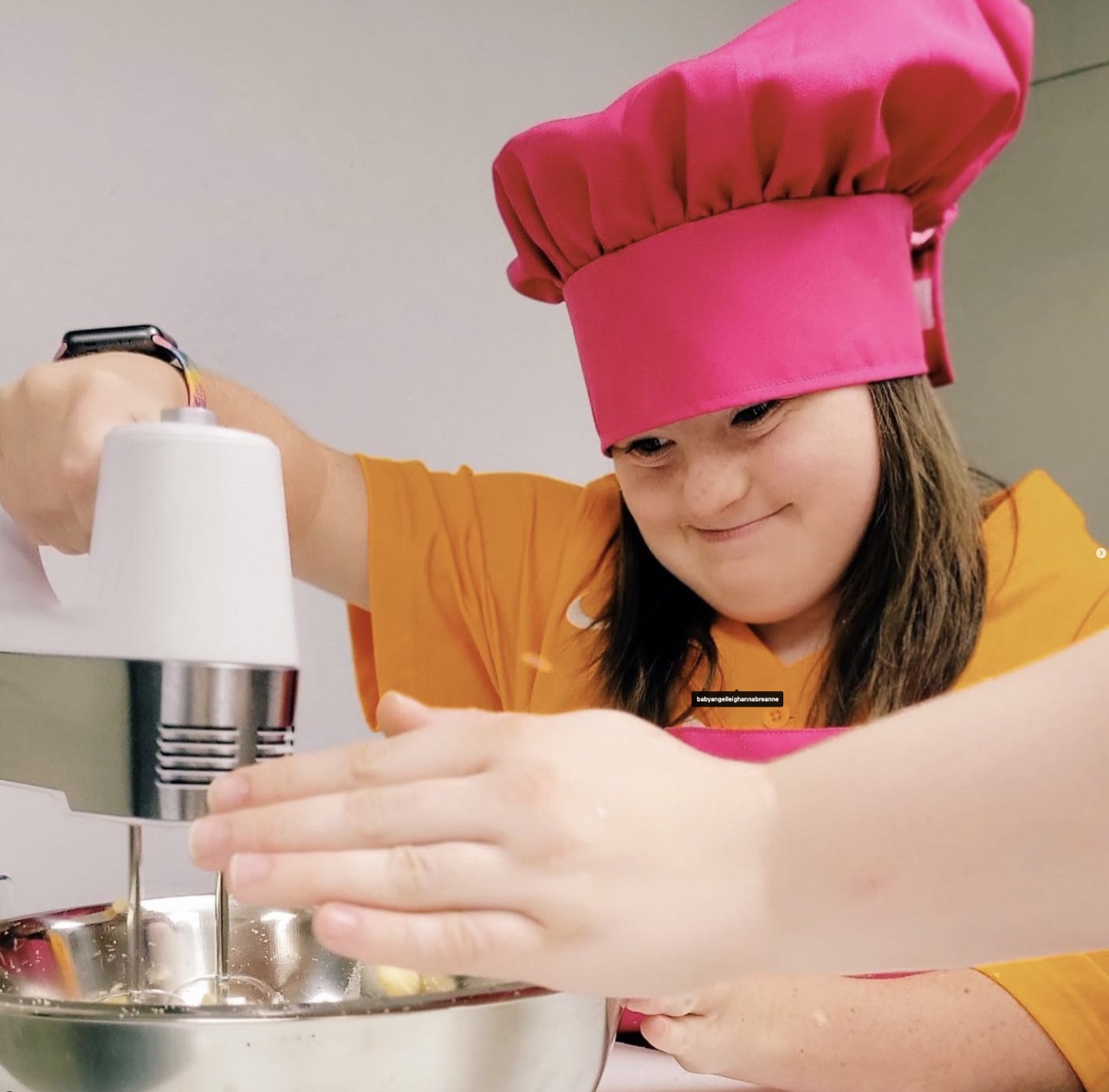 TN Believes Kitchen 1 A light-skinned young girl wearing an orange shirt and a pink chef's hat uses an electric mixer to blend ingredients in a metal bowl. She is focused on her task, and her hands are steady on the mixer.