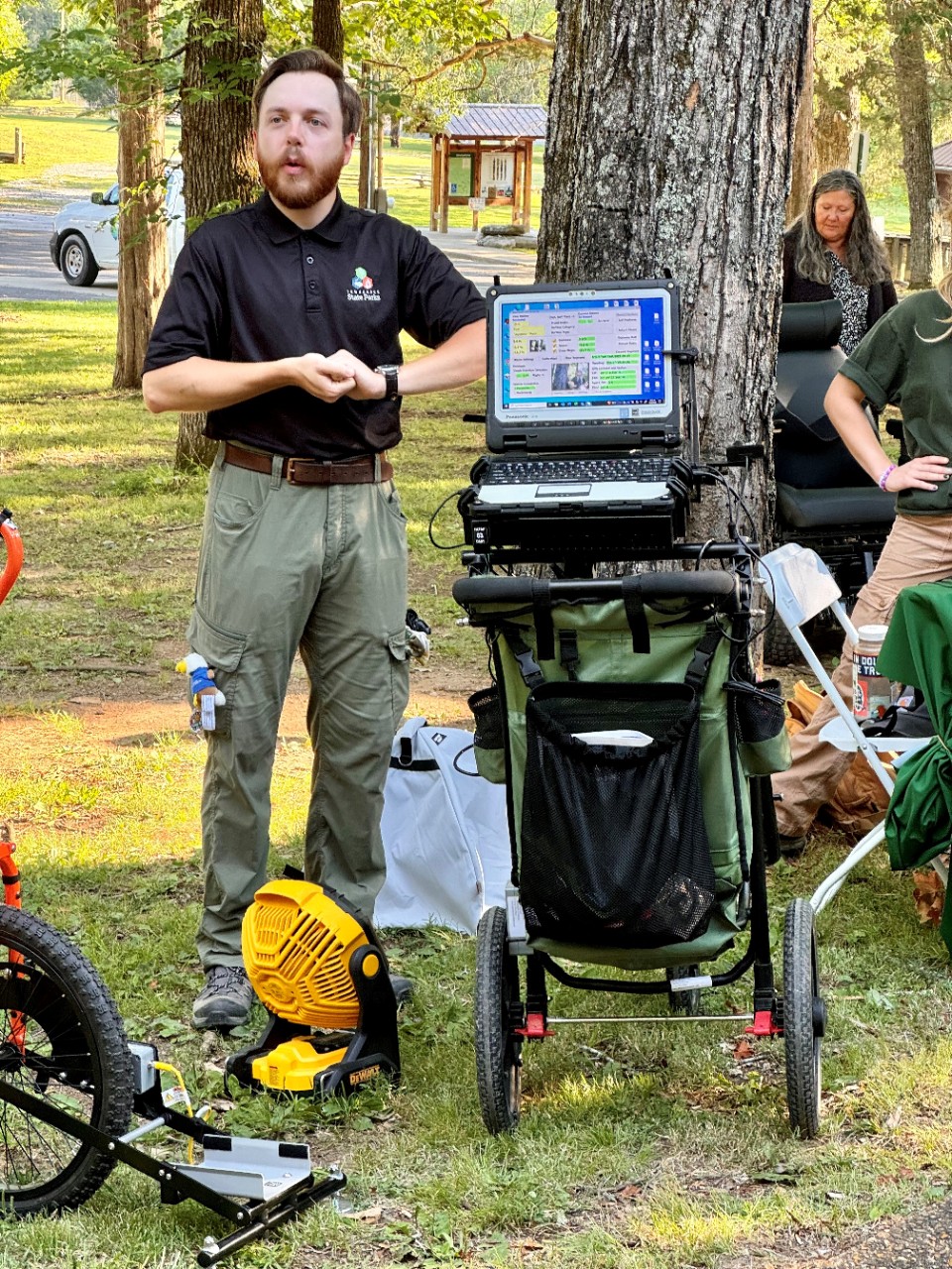 Trail assessments A State Parks employee with a light skin tone, short brown hair, and a beard, is demonstrating trail assessment equipment outdoors. He is wearing a black polo shirt with the State Parks logo and green cargo pants, standing beside a portable setup with a laptop and a pushcart equipped with specialized sensors. The demonstration is taking place in a wooded area with other park staff and visitors in the background.