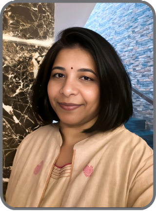 Untitled design - 1 Swathi Dasari, a woman with a medium skin tone and shoulder-length black hair, is smiling at the camera. She is wearing a light-colored top with pink embroidered details. The background features a wall with a marble design on one side and a textured stone wall on the other.