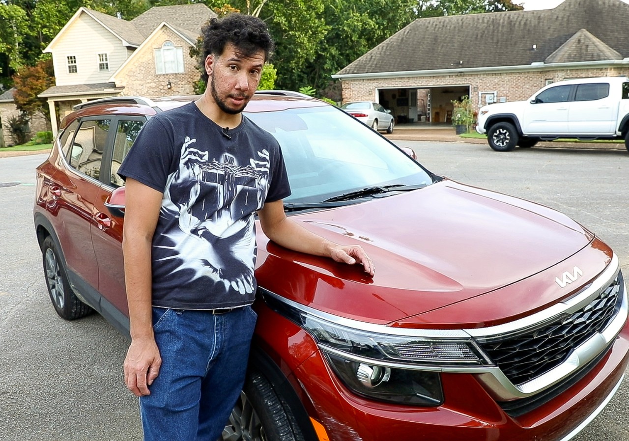 A man with a medium skin tone and short black hair is standing outside next to a red SUV. He is wearing a black t-shirt with a white graphic and blue jeans. He has one hand resting on the hood of the car and is looking at the camera. In the background, there are houses with trees around them, and a few other vehicles are parked nearby.