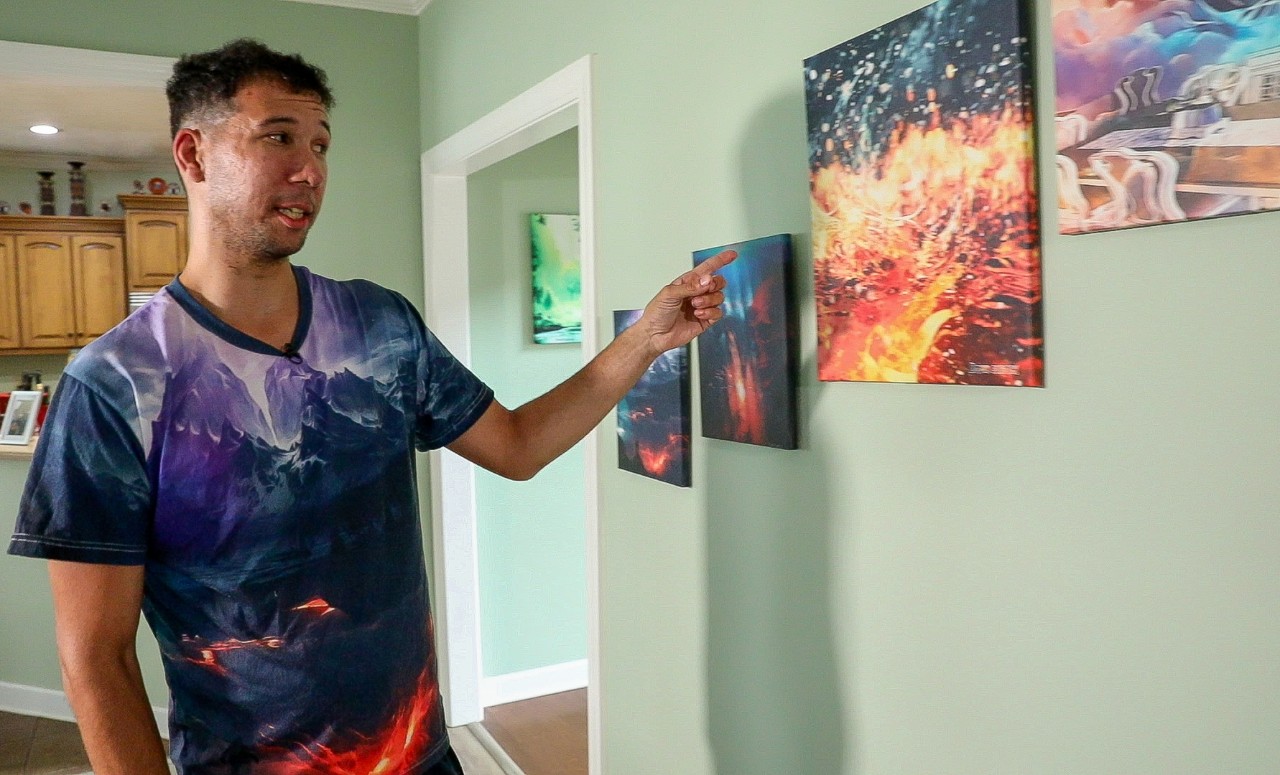 A man with a medium skin tone and short brown hair is standing in a room, pointing at a series of colorful, abstract paintings on the wall. He is wearing a tie-dye t-shirt featuring a dark mountain landscape design. The room has light green walls, and there is a doorway leading to a kitchen in the background.