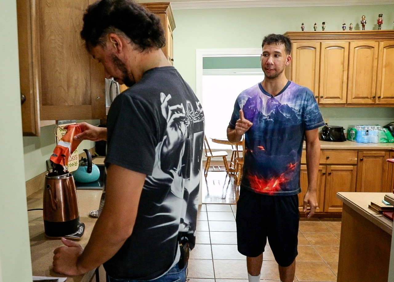 Two men with medium skin tones are in a kitchen. The man on the left, with short black hair, is facing away, pouring coffee grounds into a copper coffee maker. He is wearing a black t-shirt with a white graphic. The man on the right, wearing a tie-dye t-shirt with a mountain design and navy shorts, is standing slightly behind him, gesturing with one finger raised as if making a point. The kitchen has light green walls, wooden cabinets, and tiled floors, with a doorway leading to a dining area visible in the background.