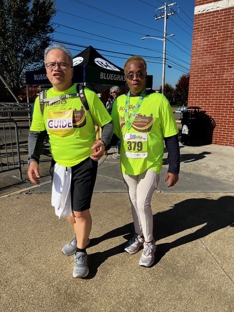 Achilles 4 Two people posing together outdoors at an event. The person on the left, a light-skinned man with short gray hair, wears a neon green "Achilles Guide" shirt, black shorts, gray sleeves, and a white towel tucked into his waistband. The person on the right, a black woman with short blond hair, wears a matching neon green "Achilles" shirt, white leggings, and sunglasses, with a race bib numbered "379" and a medal around her neck. They are standing on a sunny day in front of a red brick building and event tents.