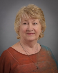 MickieWhitakerPort A portrait of Mickie Whitaker, a light-skinned woman with short, wavy blond hair and a warm smile. She is wearing a orange and teal blouse and silver jewelry, including hoop earrings and layered necklaces, one with a cross pendant. The background is a soft gray, giving a professional and polished appearance.