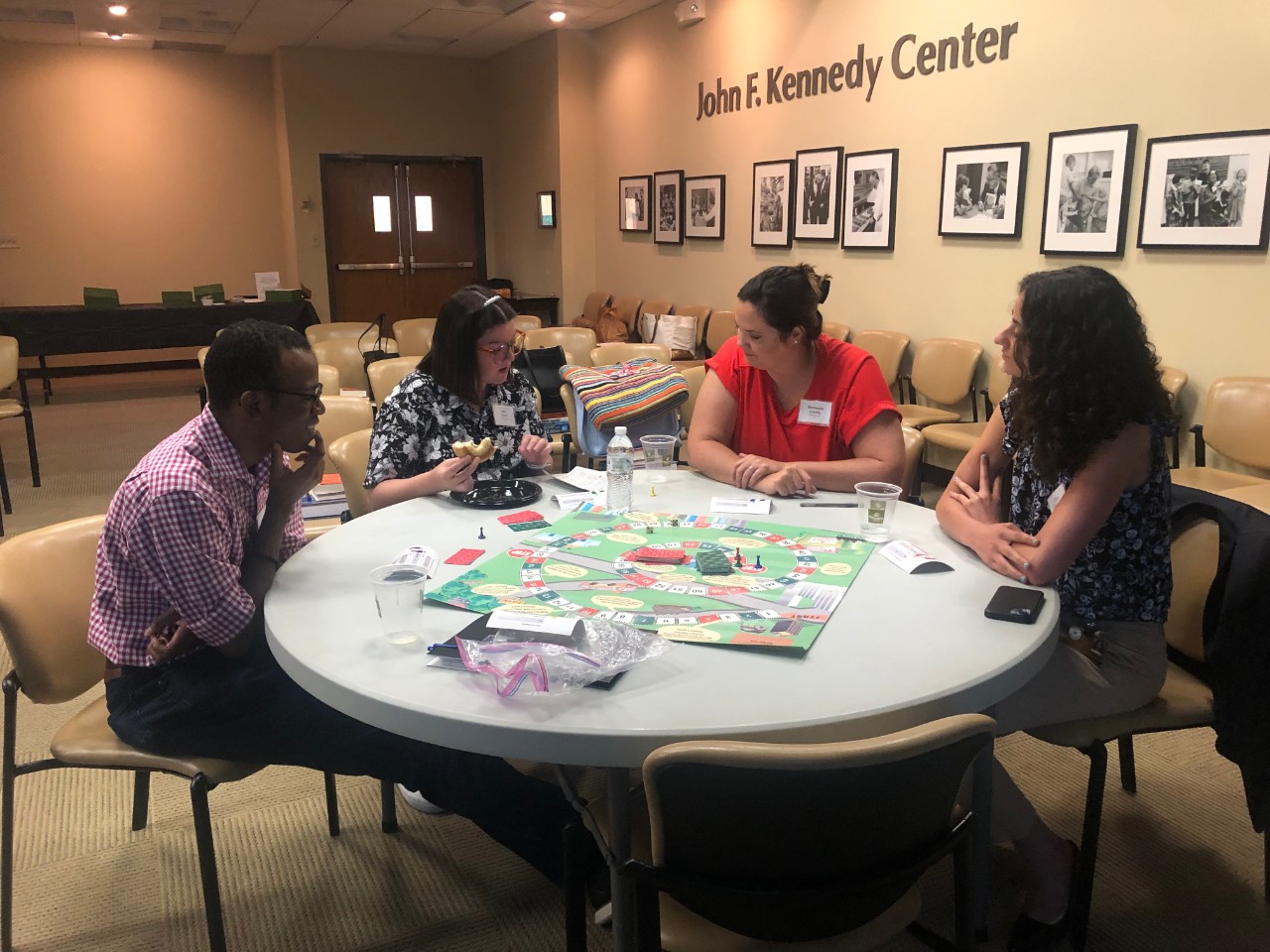 Four people sit around a circular table engaged in a board game at the John F. Kennedy Center. The group includes a man with medium brown skin wearing glasses and a pink checkered shirt, a woman with light skin and glasses eating a sandwich, a woman with light skin in a red shirt, and a woman with light skin and dark curly hair. The table has game pieces, cards, and cups, with a casual and collaborative atmosphere. The room features framed photos on the wall and beige chairs.