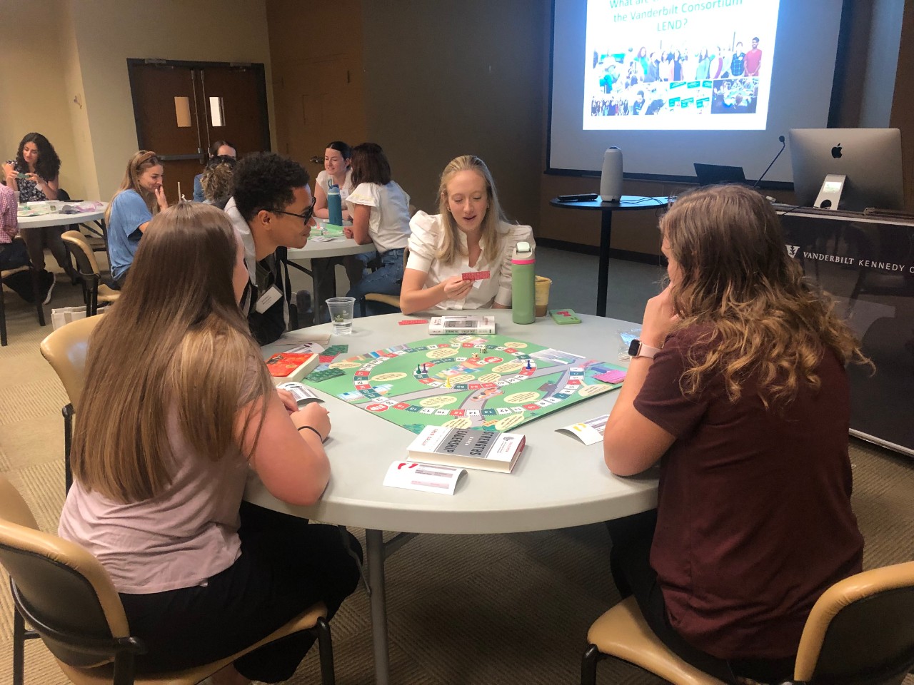 Four people sit around a table playing a board game during a workshop at the Vanderbilt Kennedy Center. The group includes a man with medium brown skin and glasses, and three women with light skin, one of whom is holding a card. The table is covered with game pieces, cards, and materials. In the background, a projector displays a slide about the Vanderbilt Consortium LEND program. Other groups can be seen at tables engaged in similar activities, in a bright and collaborative setting.