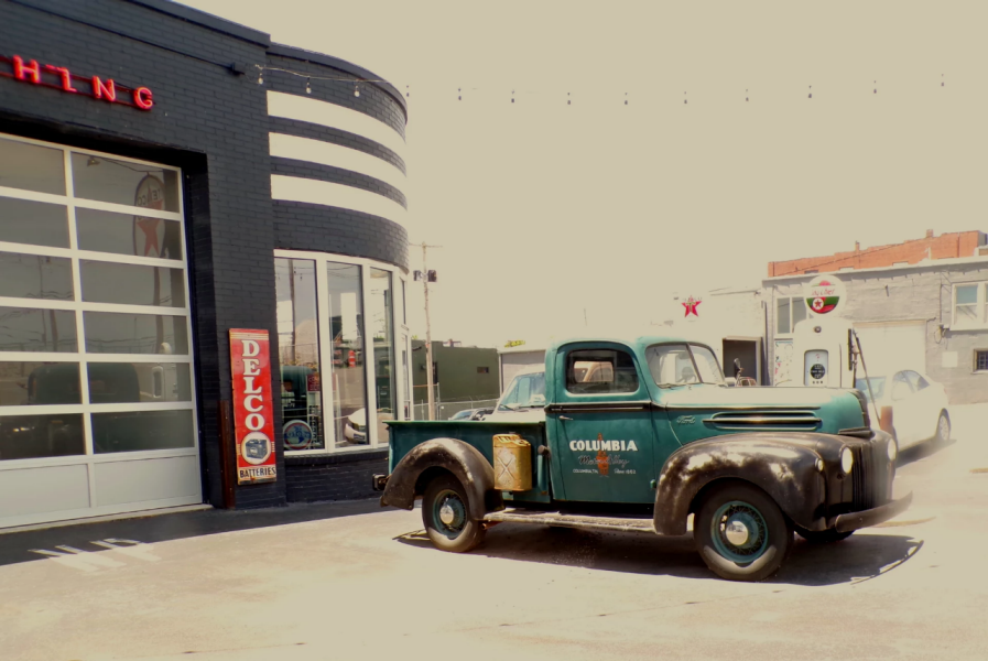Photo 1 A vintage green Ford pickup truck with rusted fenders is parked outside an old-fashioned gas station. The truck has "Columbia" and other faded lettering on its door, giving it a nostalgic, well-worn look. The gas station has a black brick exterior with curved white stripes, large glass windows, and a red "Delco Batteries" sign mounted beside the garage door. A retro gas pump with a red Texaco star logo stands in the background, along with a white car and an industrial-style building. The bright, slightly overexposed lighting gives the scene a vintage feel.