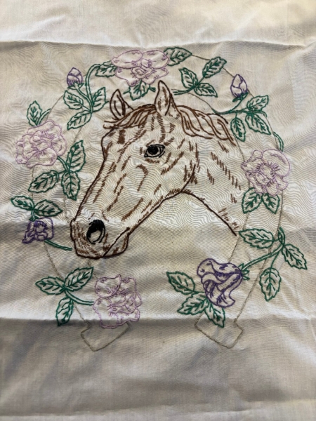 Shoe Horse An embroidered design on white fabric features a detailed horse's head outlined in brown thread, with expressive eyes and shading for depth. Surrounding the horse is a circular wreath of green leaves and delicate purple flowers, stitched with intricate detail. Below the horse, faintly outlined objects resembling a ribbon or banner are visible. The fabric has some wrinkles, giving it a handmade, in-progress feel.