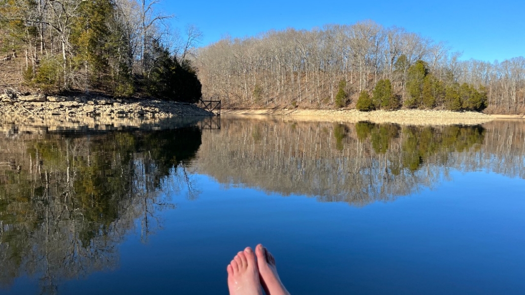 A serene lakeside scene shows a pair of bare feet in the foreground, resting above the calm water of Tims Ford Lake. The lake reflects the surrounding landscape of leafless trees, evergreens, and rocky shoreline, creating a near-perfect mirror image. The sky is clear and blue, with sunlight casting a warm glow on the trees and rocks. A small wooden dock is visible in the distance. 