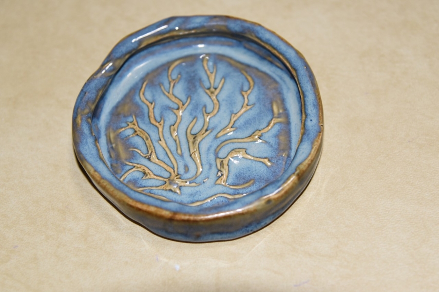 A handmade ceramic coaster that features a light blue glaze with earthy brown accents. At its center, a raised branching design resembling flames or a tree extends outward, with the unglazed ridges contrasting against the glossy surface. The edges of the coaster are slightly irregular, adding to its organic, handcrafted feel.