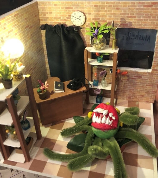 A miniature diorama of a flower shop with a brick wall interior, shelves filled with tiny vases and plants, and a wooden desk with a black telephone and books. A large, carnivorous plant with a red, toothy mouth and fuzzy green tentacle-like vines sits at the center, resembling Audrey II from Little Shop of Horrors. A black curtain covers part of the back wall, and a clock hangs above it. The window displays the shop's name, partially readable as "Mushnik’s Flower Shop."