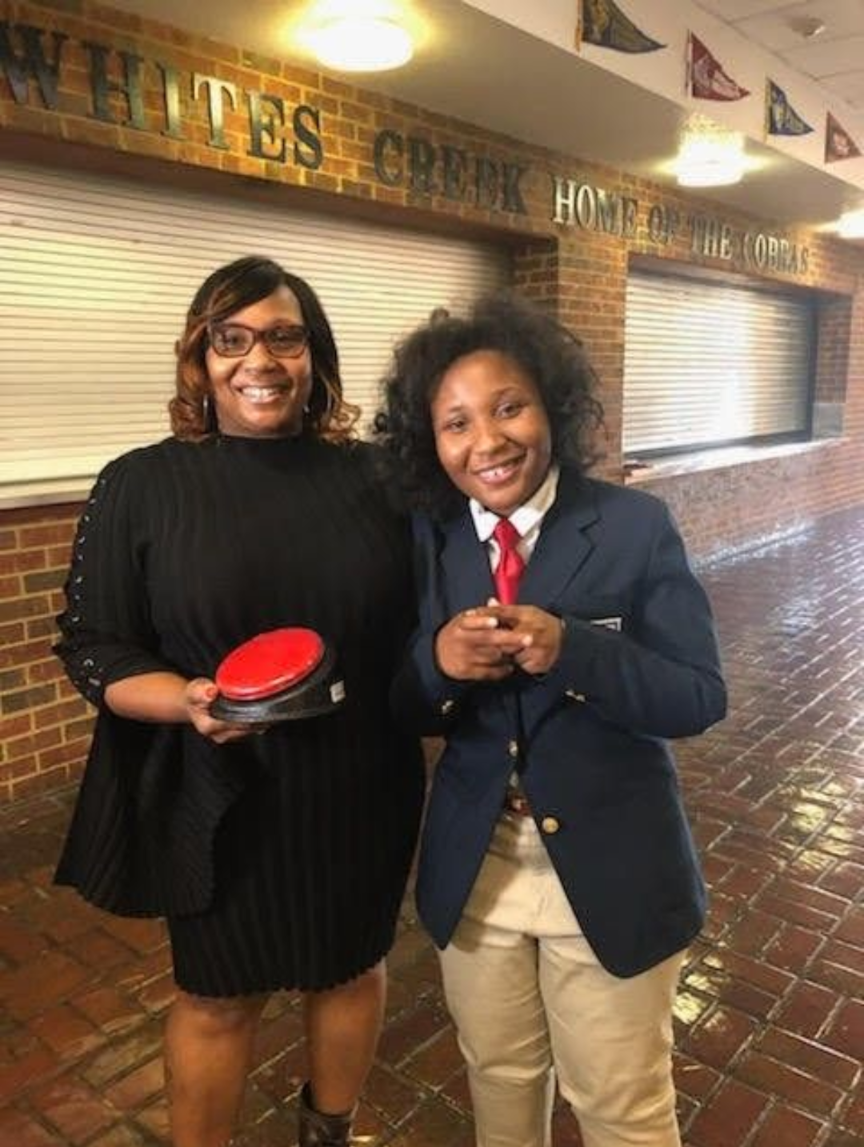 A photograph of Tamara Tuckson and McKenzie Tuckson, both African-American, standing together in a school hallway. Tamara, on the left, has medium brown skin, wears glasses, has shoulder-length wavy brown hair, and is dressed in a black pleated dress with decorative buttons on the sleeves. She holds a red communication button device called a Big Mack switch. McKenzie, on the right, has medium brown skin and curly black hair. She  is dressed in a navy blue blazer with gold buttons, a white shirt, a red tie, and beige pants. She is smiling and holding her hands together in front of her. Both are smiling warmly at the camera.