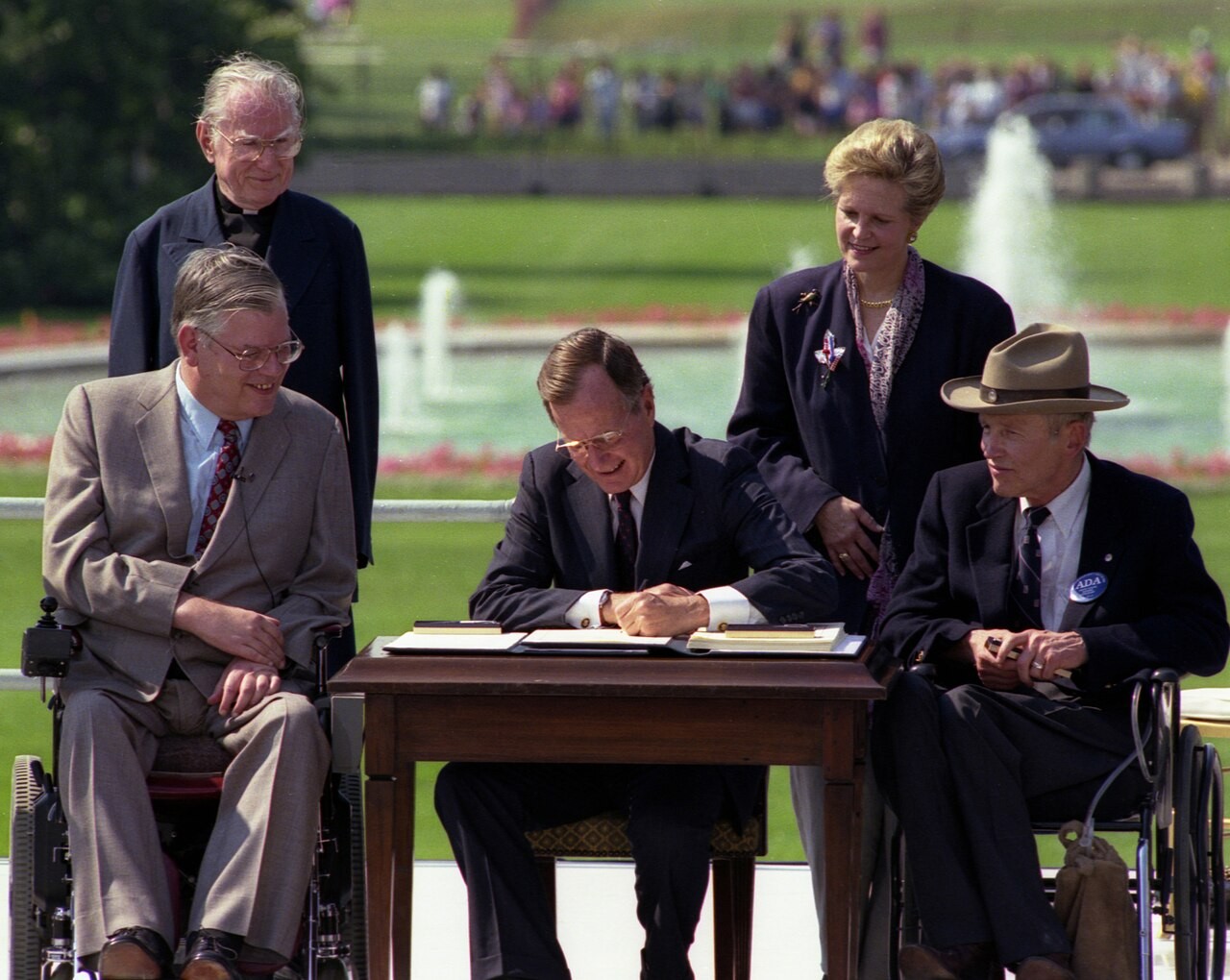 President George H. W. Bush, a white man in a dark suit, is seated at a table signing the Americans with Disabilities Act in an outdoor ceremony on the White House lawn. Surrounding him are five disability rights advocates, including two white men in wheelchairs. One man wears a tan suit and red tie; the other wears a dark suit and a cowboy hat with an ADA button. A woman and another man stand behind them, smiling. A fountain and crowd are visible in the background.