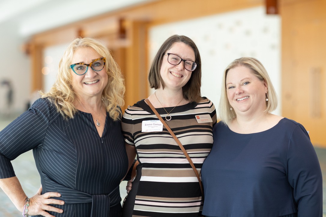 Three women stand smiling together at the 2024 Fall Disability Rights TN Disability Employment Awareness Luncheon. Lisa has long curly blonde hair and wears glasses and a dark pinstripe dress. Emma, with a name tag, has shoulder-length brown hair, glasses, and a striped dress with a crossbody bag. Anna has straight blonde hair and wears a navy blue top. All have light skin tones.