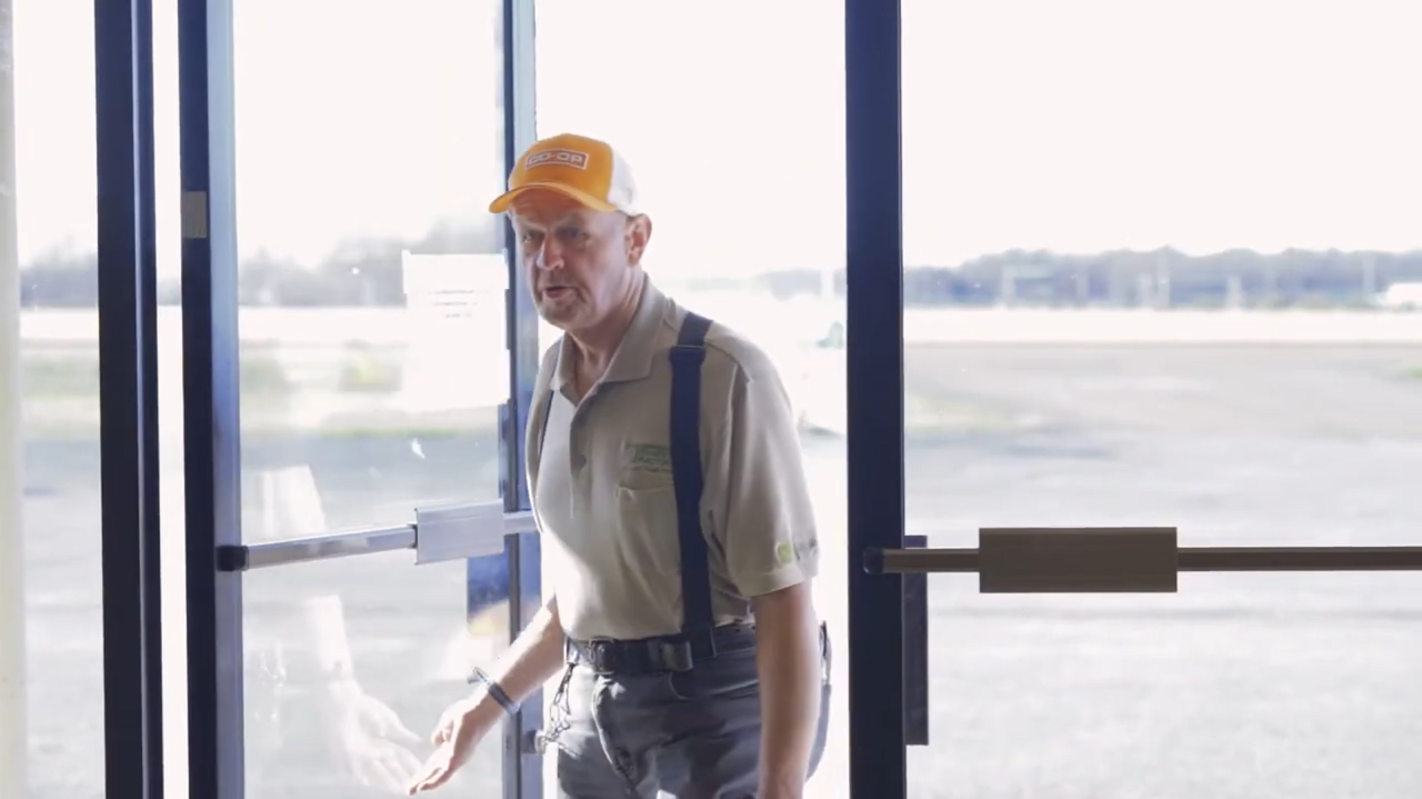danny entering store A fair-skinned older man, Danny Simmons, wearing a beige Tennessee Tractor polo, blue suspenders, and an orange and white CO-OP cap, is entering a building through a glass door with bright sunlight streaming in behind him.