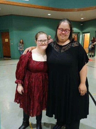 jackie at dance At a Valentine’s Dance, Jackie Taylor (right) wears a black dress with lace detail and pink glasses. She stands with a friend who has light skin and wears a red patterned dress with black boots. The two smile together in a gymnasium-style venue, enjoying the evening.