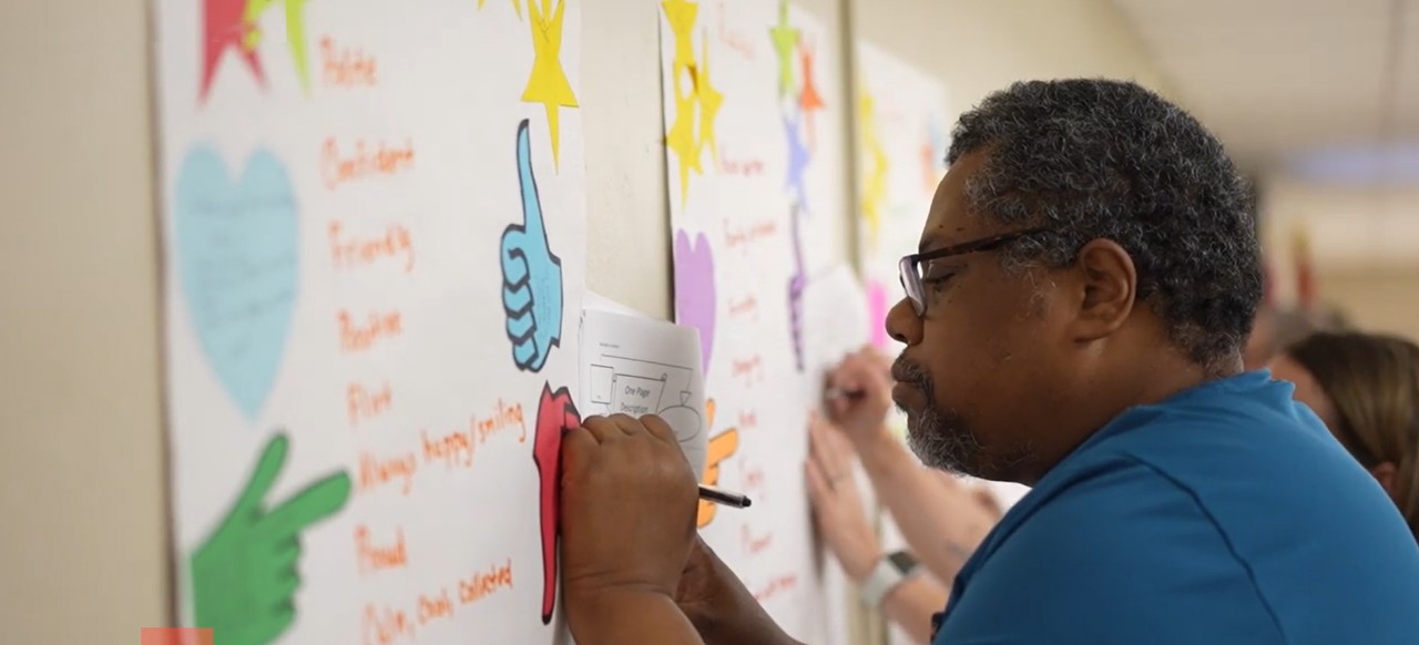 A man with dark skin, glasses, and black-and-gray curly hair and partial beard writes on a paper held against a brightly colored poster on a wall. 
