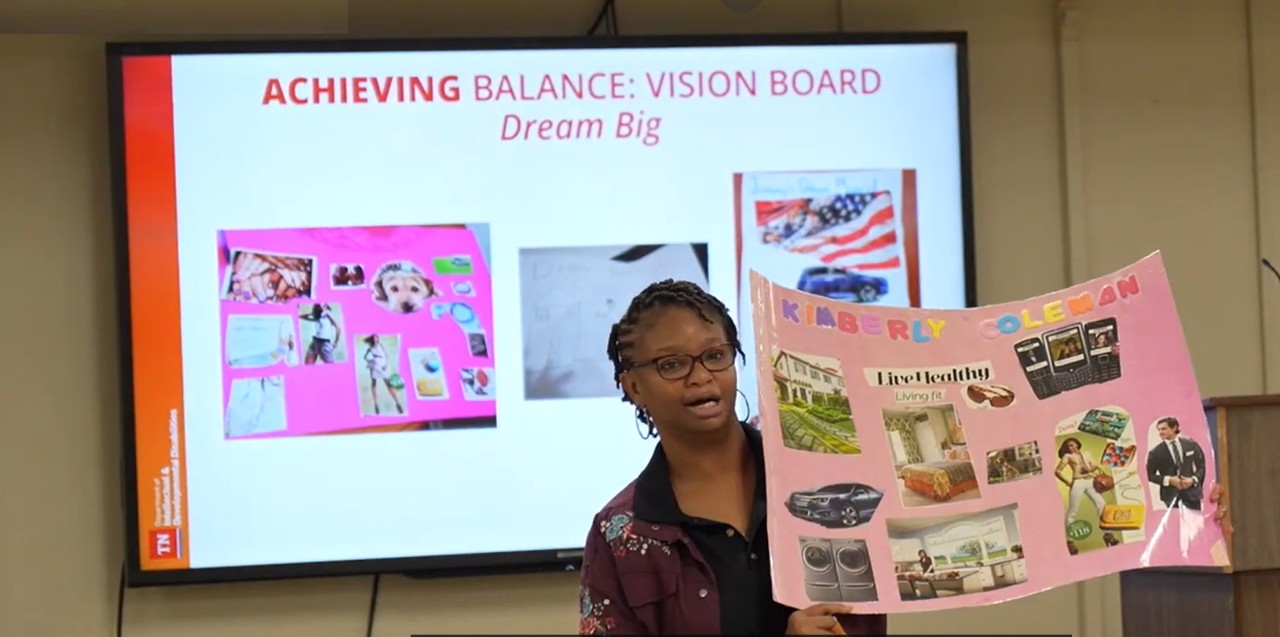 A young woman with dark skin, glasses, hoop earrings, and short cornrow braids holds up a pink poster that reads “KIMBERLY COLEMAN” and features pictures from magazines of fashion, a car, a home, mobile devices, and more. A screen is visible behind her with text that reads “ACHIEVING BALANCE: VISION BOARD – Dream Big”