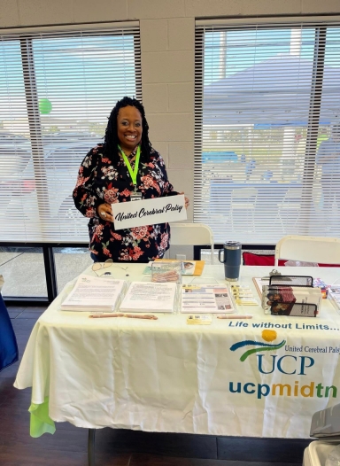 A woman with dark skin and long, braided hair is wearing a flowered shirt and conference badge and stands behind an exhibit table with materials. The woman holds a sign that reads, “United Cerebral Palsy.” 