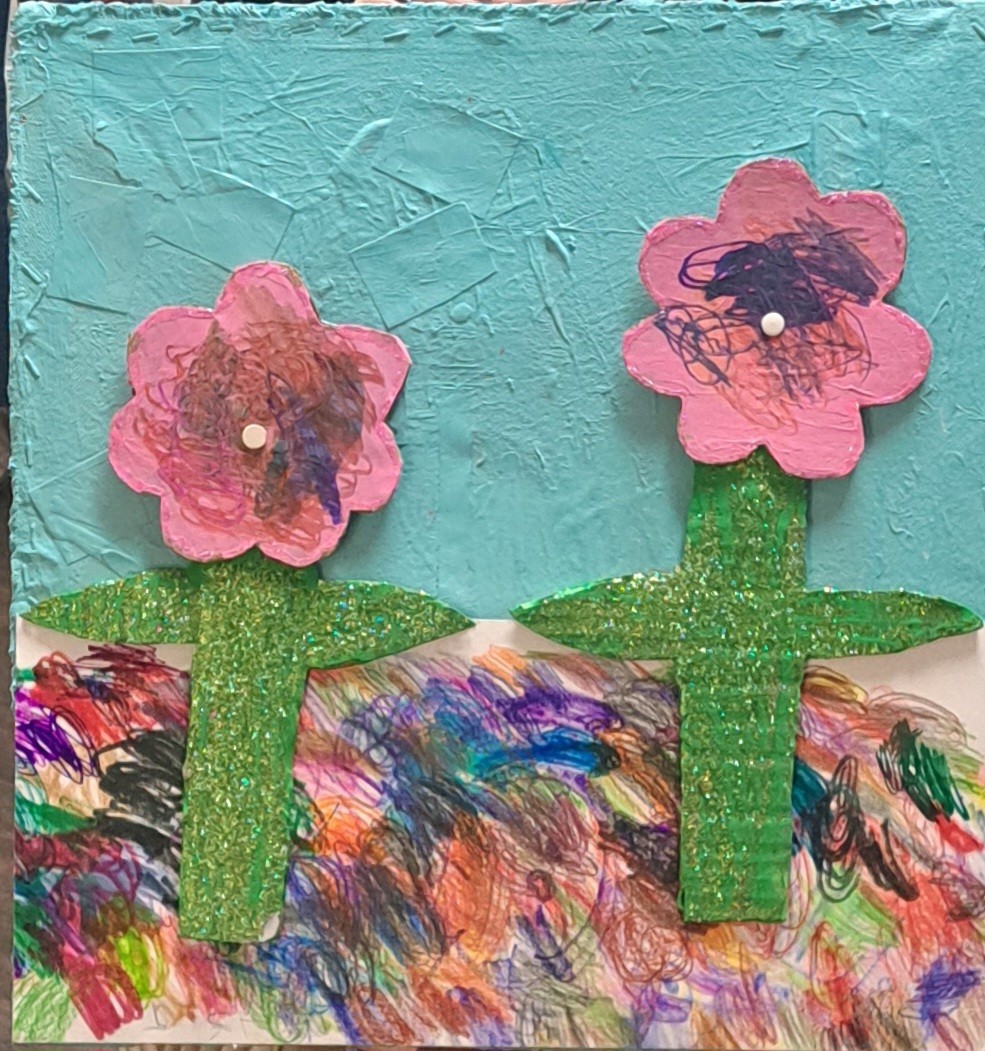 A mixed media artwork shows two simple flower shapes standing side by side against a light blue background. The flowers have pink petals with scribbled centers, and green glitter stems that stand out from the surface. Below them, a strip of colorful crayon marks creates a messy, layered ground with bright, overlapping colors.