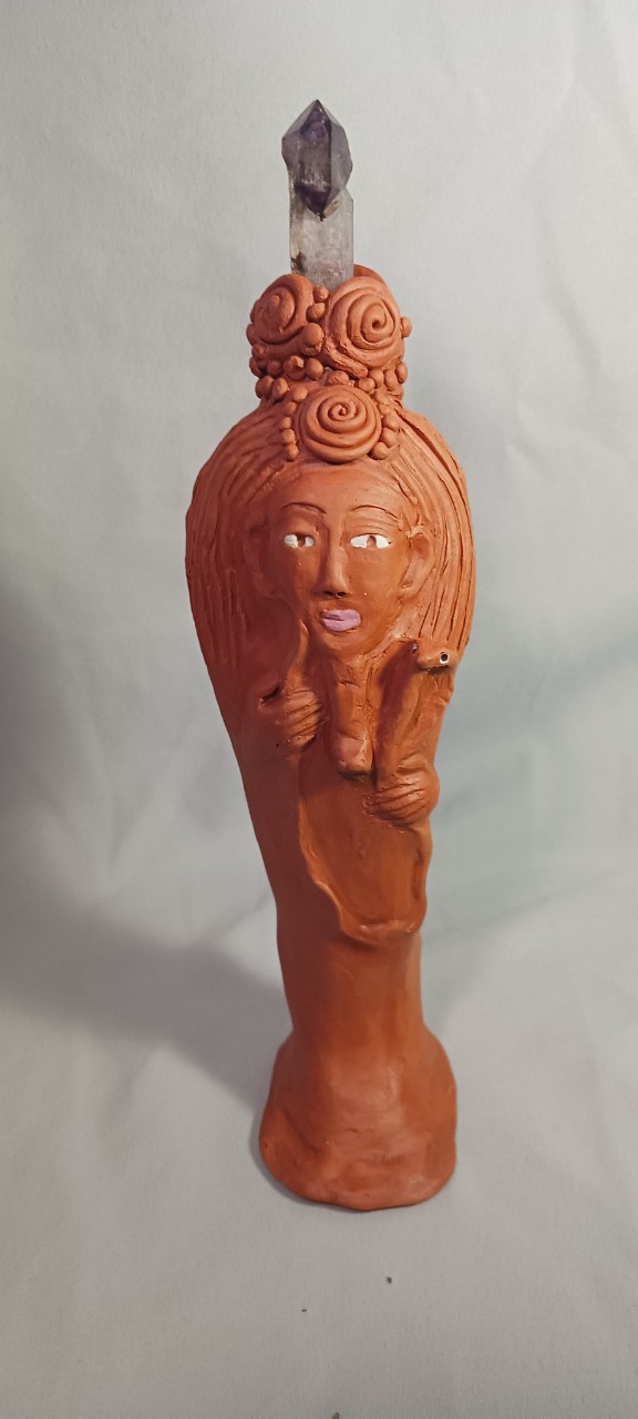 A clay sculpture shows a tall, narrow figure with a human face and long neck, standing upright against a plain background. The figure has reddish-brown clay skin, carved facial features, and textured hair with spiral shapes on top, holding a clear and purple colored crystal that ends in a point. The surface looks hand-shaped, with visible marks and a matte, earthy finish.