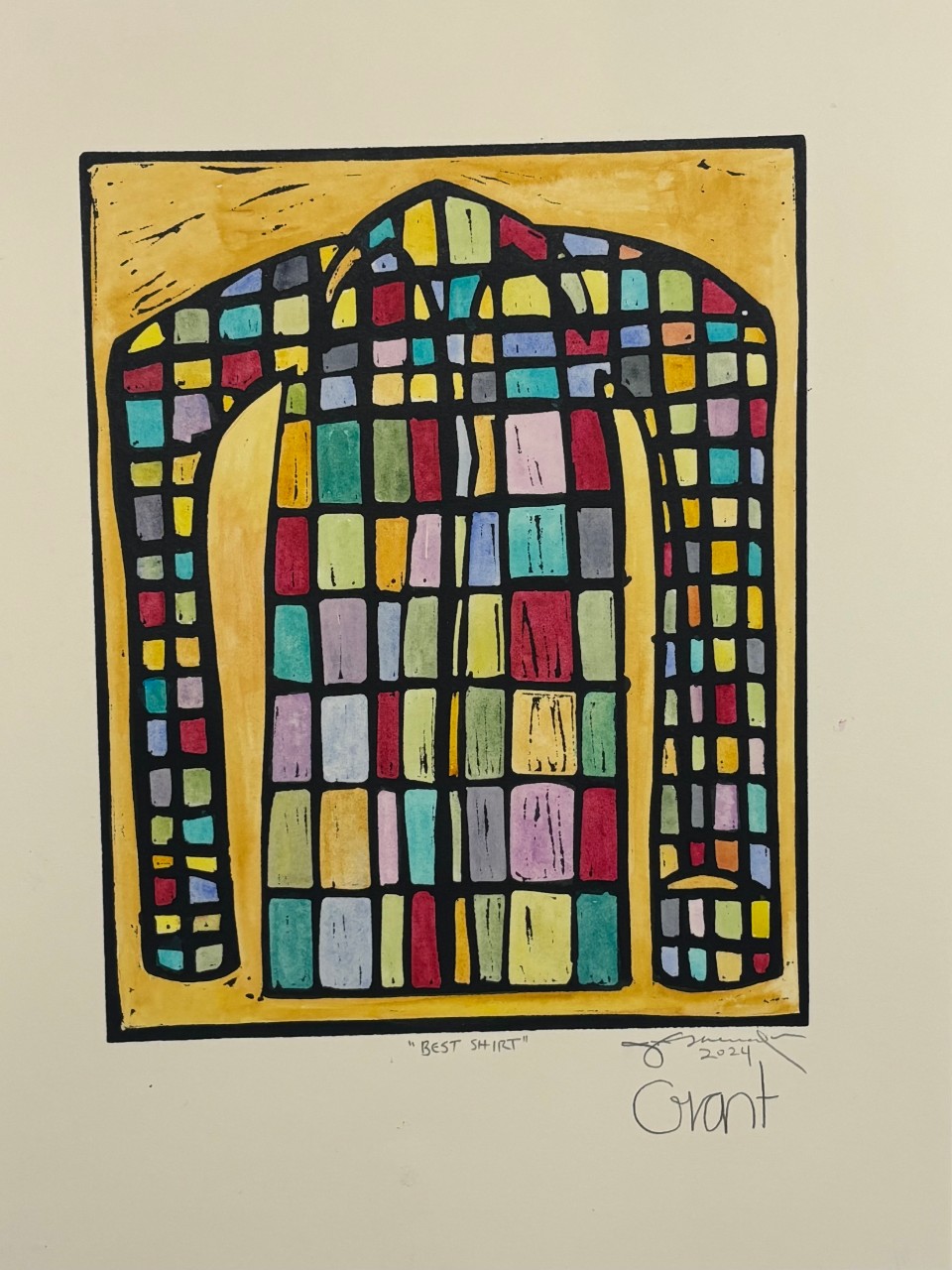 A painting shows a shirt shape outlined in black, filled with small blocks of many colors like a grid. The background is a warm yellow, which makes the colors inside the shirt stand out. The paint has visible brush marks and created simple, bold shapes.