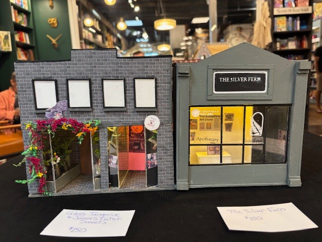 A miniature model shows two storefronts side by side, viewed straight from the front. One building has a gray brick face with bright flowers around the doorway, and the other has a sign reading “The Silver Fern” above a glowing front window. Small details in the doors, windows, and trim give the piece a careful, handmade look.