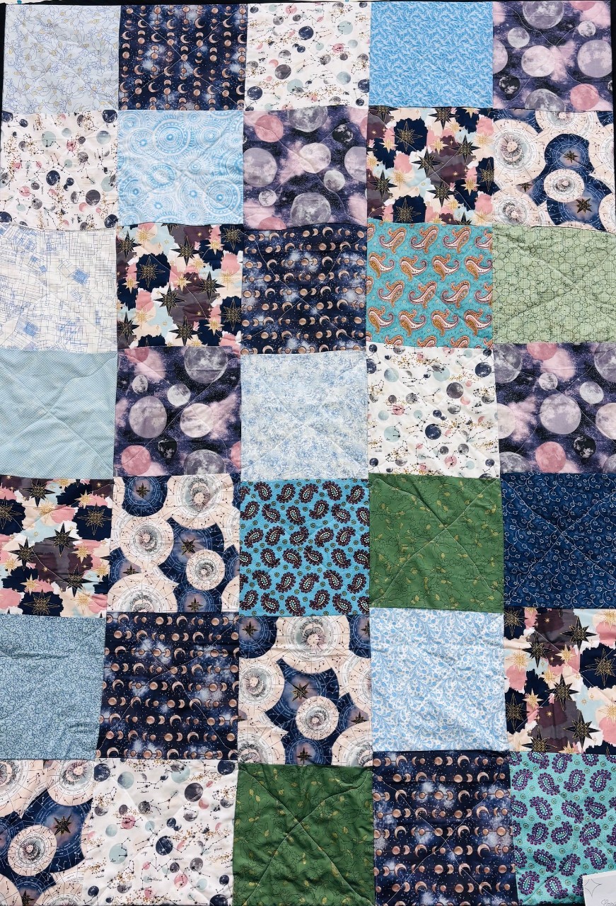 A patchwork quilt is made from square fabric blocks arranged in a neat grid. Most of the colors are cool blues, purples, teals, and soft greens, with patterns that include moons, stars, circles, paisleys, and small floral designs. Seams and stitched lines create a soft quilted texture across the whole piece.