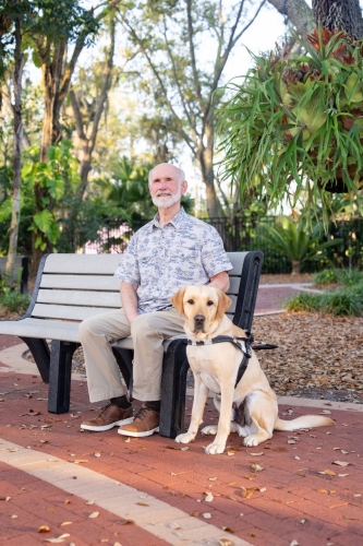 Mike Harrell sits on a park bench with his guide dog, Simba, seated beside him. Mike wears a light patterned shirt and khaki pants, while Simba, a yellow Labrador, wears a black guide harness. Trees, plants, and a brick walkway surround them in a sunny outdoor setting.
