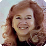Smiling woman with light skin and long wavy reddish-blonde hair, wearing a dark top, in front of a light-toned background.