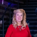 Opening note - Lori Kissinger Smiling woman with light skin and short brown hair, wearing a black blazer, pinstriped dress, and pearl necklace, standing indoors.