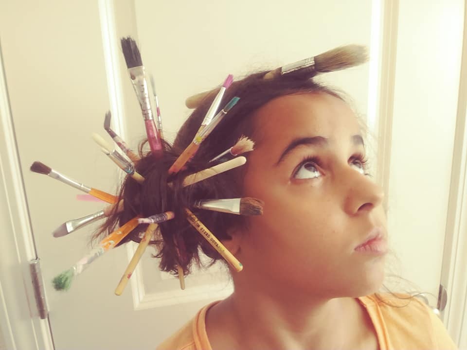 a close-up photo of a young child in a yellow shirt staring up at the ceiling. The child’s hair is dark brown, and there is an array of paintbrushes woven throughout the hair