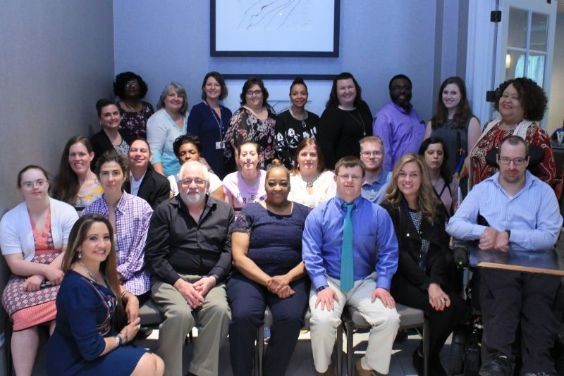 97 Partners 2018-19 graduation group photo group photo of a diverse group of about 30 folks with and without different types of disabilities, posing outside a conference room in a sunlit hallway