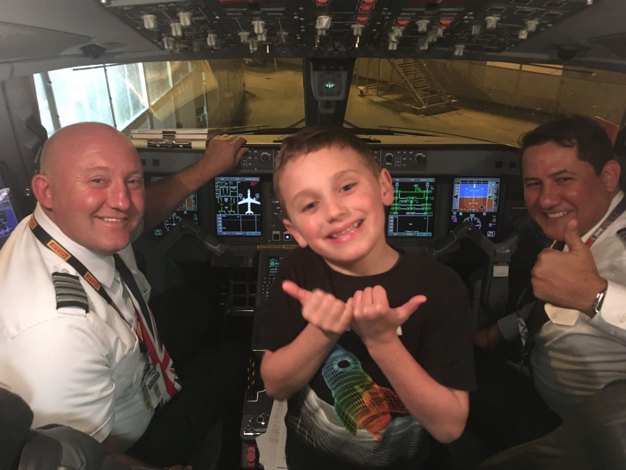 97 Fly the Friendly Skies pic the cockpit of a commercial airplane. There are two pilots smiling at the camera; one is giving a “thumbs up” sign. In the middle is a young boy, Jessica Gant’s son. He is smiling and giving two “thumbs up”.