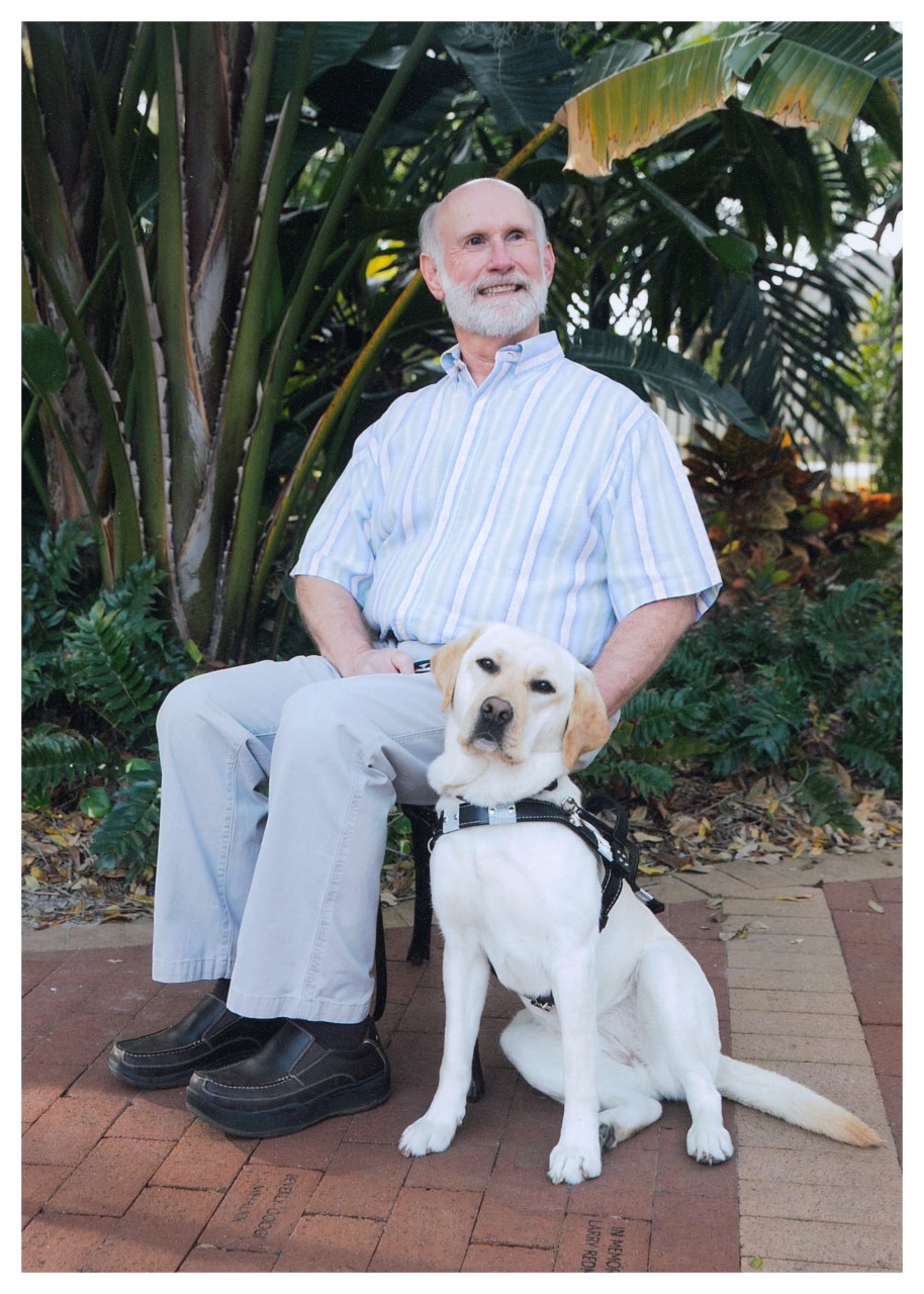 97 Mike Harrell and Honor II a gentleman with white hair and a beard is wearing a striped button up shirt and nice pants, sitting in a chair; next to him on the ground is a golden dog wearing a service dog vest