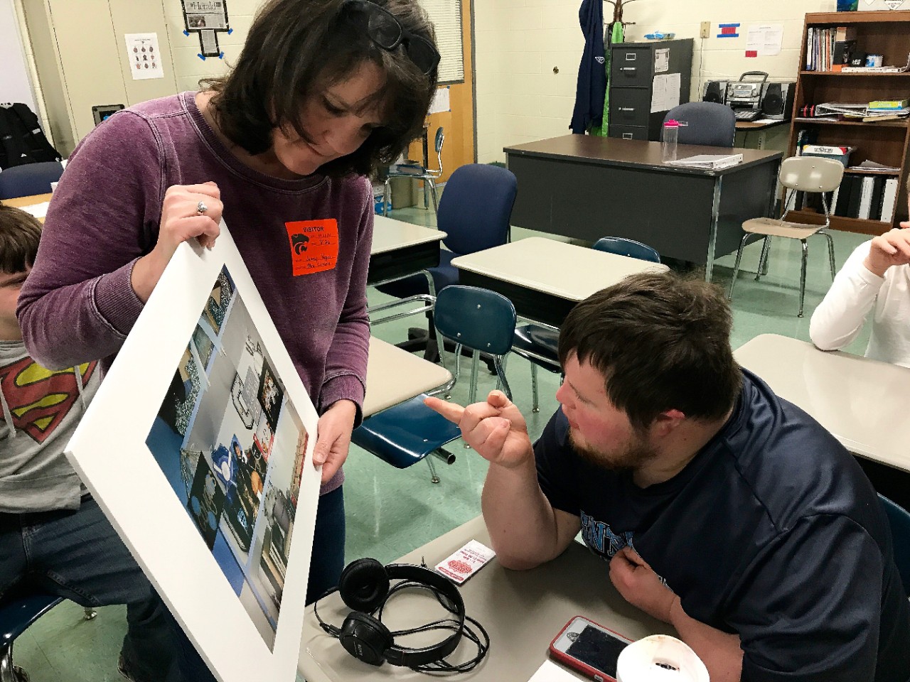98 AbleVoices 3 Author of the article Jen Vogus holds up a framed photo in a classroom and the young adult student seated in front of her is leaning forward and pointing at it
