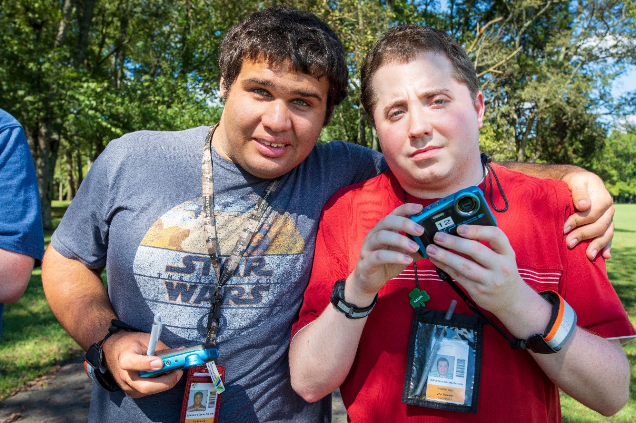 98 AbleVoices 5 two young men with disabilities stand together with their arms around each other and holding small cameras during an AbleVoices activity outside