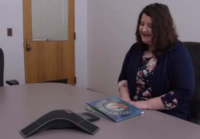 a woman sits in a room with a children's book on a table in front of her; she is speaking into a conference phone speaker