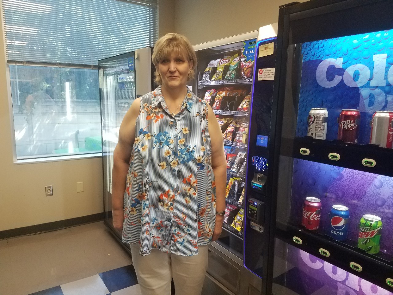 98 Sharon Paris-Treadway a blonde woman in a flowery sleeveless shirt stands in the break room and cafeteria of the state office building where she works, with vending machines behind her