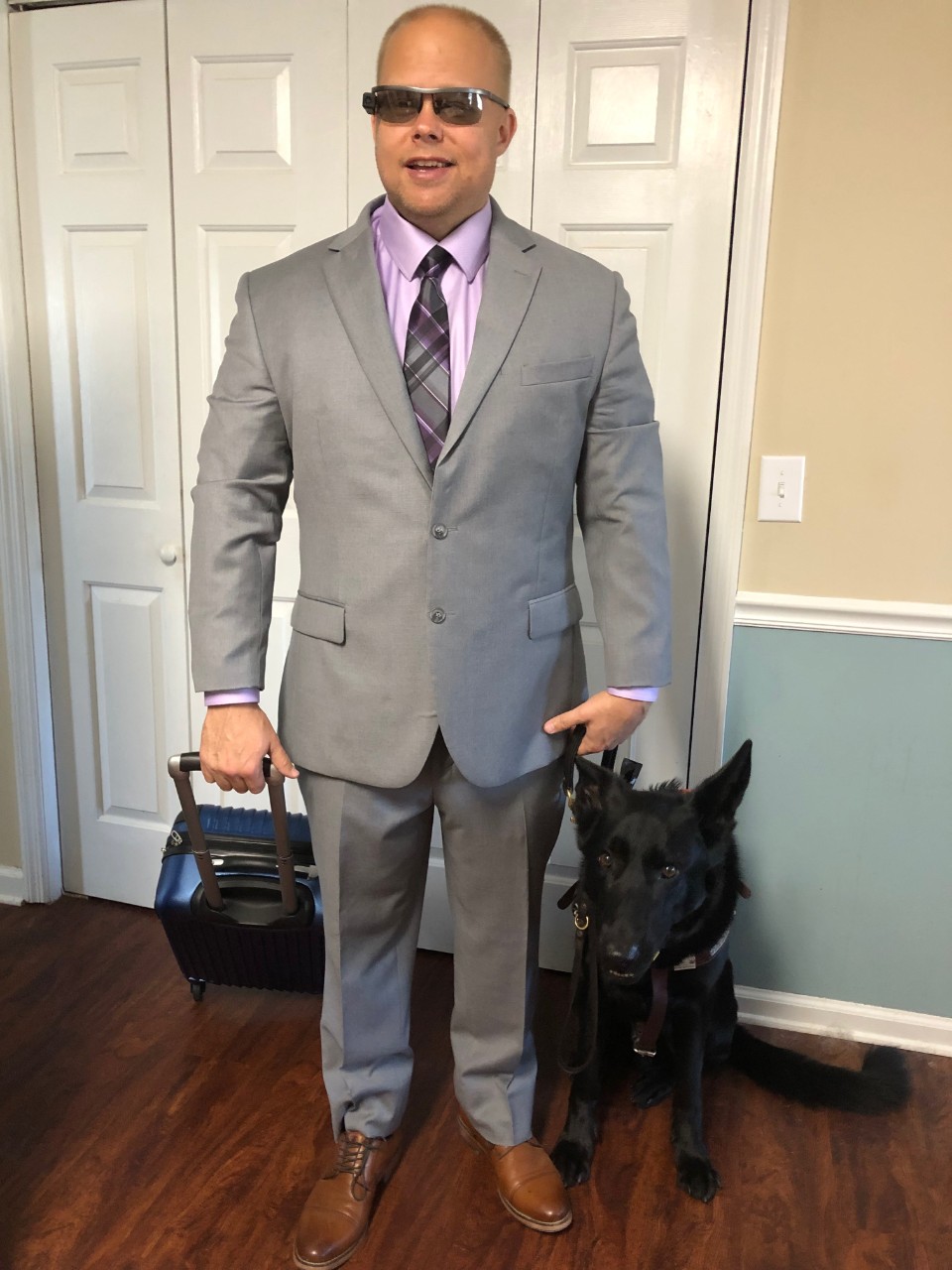 BG99 James Brown and dog with google glasses picture of article author James Brown standing in front of a closet door with his black assistant dog, Vantis. James is very professionally dressed in a gray business suit, and he is pulling a blue suitcase on wheels. His other hand holds his dog’s harness.