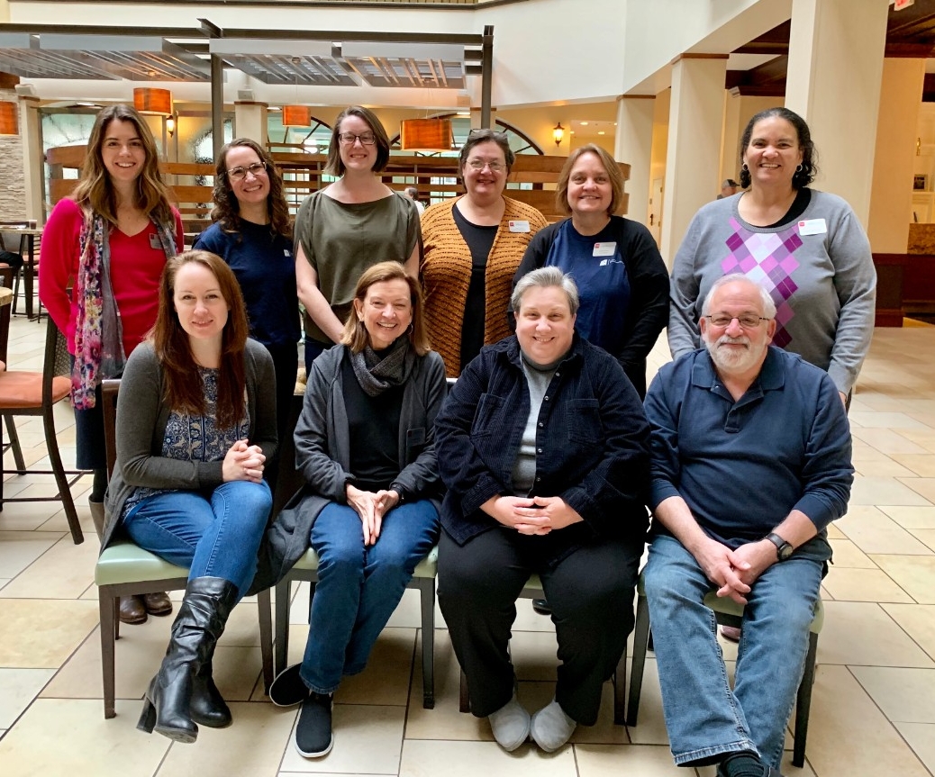 Staff 2019 retreat a group of the Council staff - 9 women and Ned, seated and standing in 2 rows in the lobby of the hotel where the Council meets