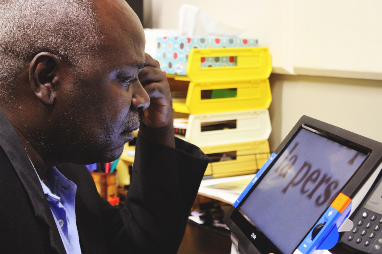BG99 Ernest using a CCTV, which enlarges text An African gentleman with white hair and a black suit jacket looks at a CCTV with enlarged text at a desk