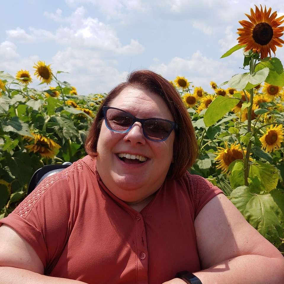 BG99 Suzanne Colsey a close-up picture of the article author, Suzanne Colsey, sitting in a field of tall, bright sunflowers. Suzanne is wearing a coral-colored blouse and smiling. The caption reads, “Suzanne Colsey enjoying a beautiful day.”