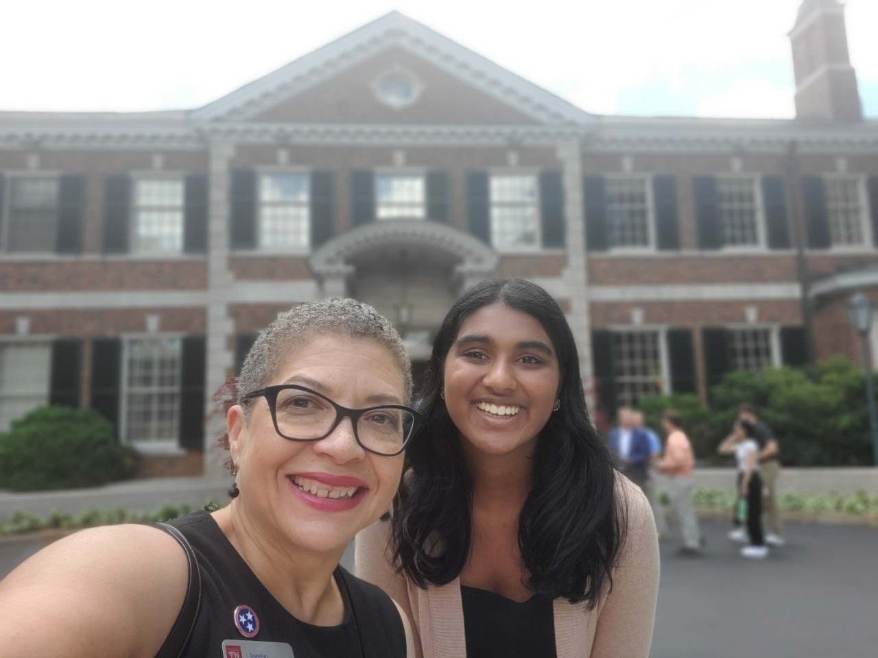 0e3f364a-9971-1872-ad84-d7f8a75659ad Selfie of two smiling women in front of a brick university building. Left: older woman with short curly hair, glasses, wearing black top with TN flag pin. Right: younger woman with long dark hair, wearing light cardigan. Background shows colonial-style architecture and people walking.