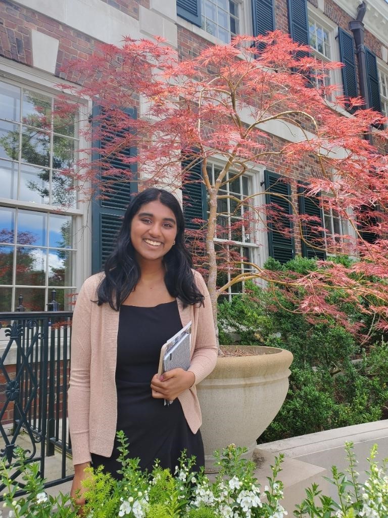 2ba72eb9-ca79-2223-0987-089dddfc0c0d Eshani, who is a young woman, smiling at the camera. Eshani has long dark hair and brown skin. She is wearing a black top with a light pink cardigan and is holding what appears to be a notebook or folder. Eshani is standing in front of a brick building with large windows and black shutters, with a vibrant red Japanese maple tree in full foliage behind her.
