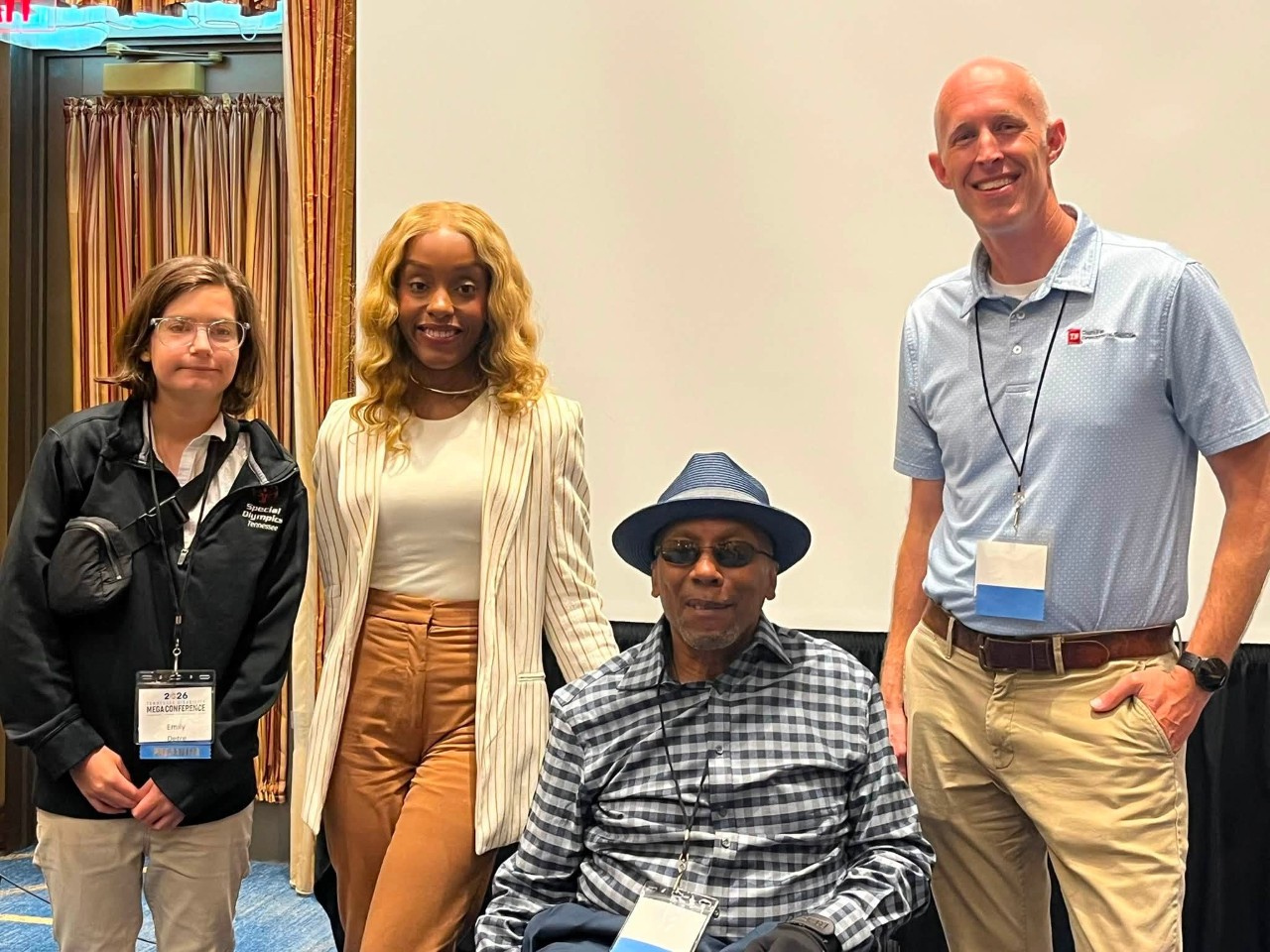 782544602_1519651486864482_1332262618621088570_n Four people posing together at in a conference session room - three standing and one seated in a wheelchair, all smiling at the camera.