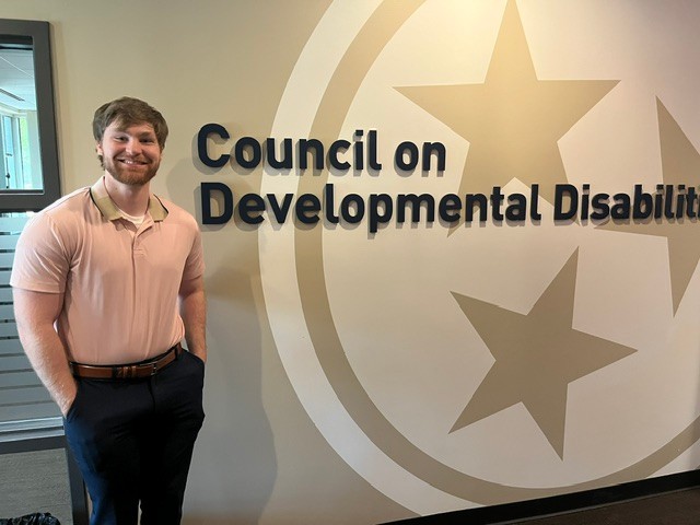 e1a9339f-ac4d-4a0b-bf07-0d3a8e86c4dc Keith Dear, a smiling young man with short brown hair and a beard in a pink button-down shirt, stands in front of a wall bearing the gold Tennessee tri-star emblem and the words "Council on Developmental Disabilities."