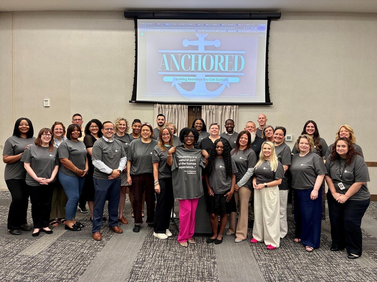 tshirts tlads About 30 people stand together smiling in a conference room, most wearing matching gray T-shirts. One person in the center holds up a shirt with the words "Disability is a natural part of the human experience..." from The Developmental Disabilities Act. A projection screen behind them reads "ANCHORED: Building Resilience You Can Sustain."