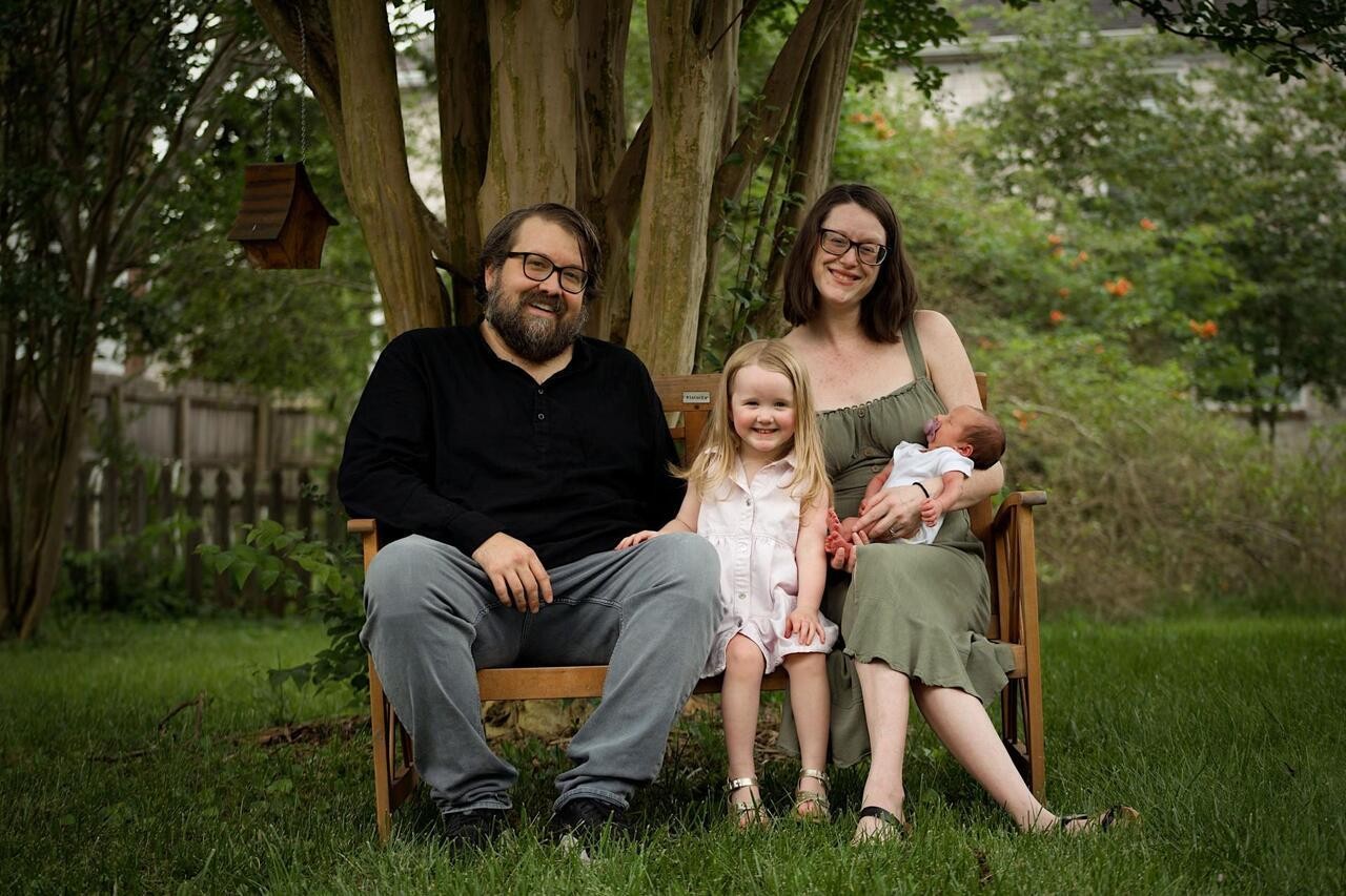june 2 Emma and TJ sit on a wooden bench outdoors with Bonnie and baby Jesse. Bonnie sits between them, smiling at the camera, while Emma holds Jesse in her arms. The family is surrounded by green grass, trees, and garden plants.